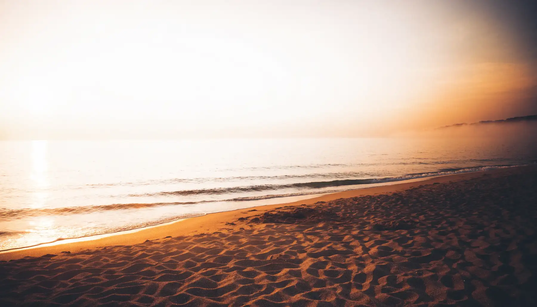 Orilla de una playa al amanecer: arena, olas suaves y una luz naranja que se funde con la bruma